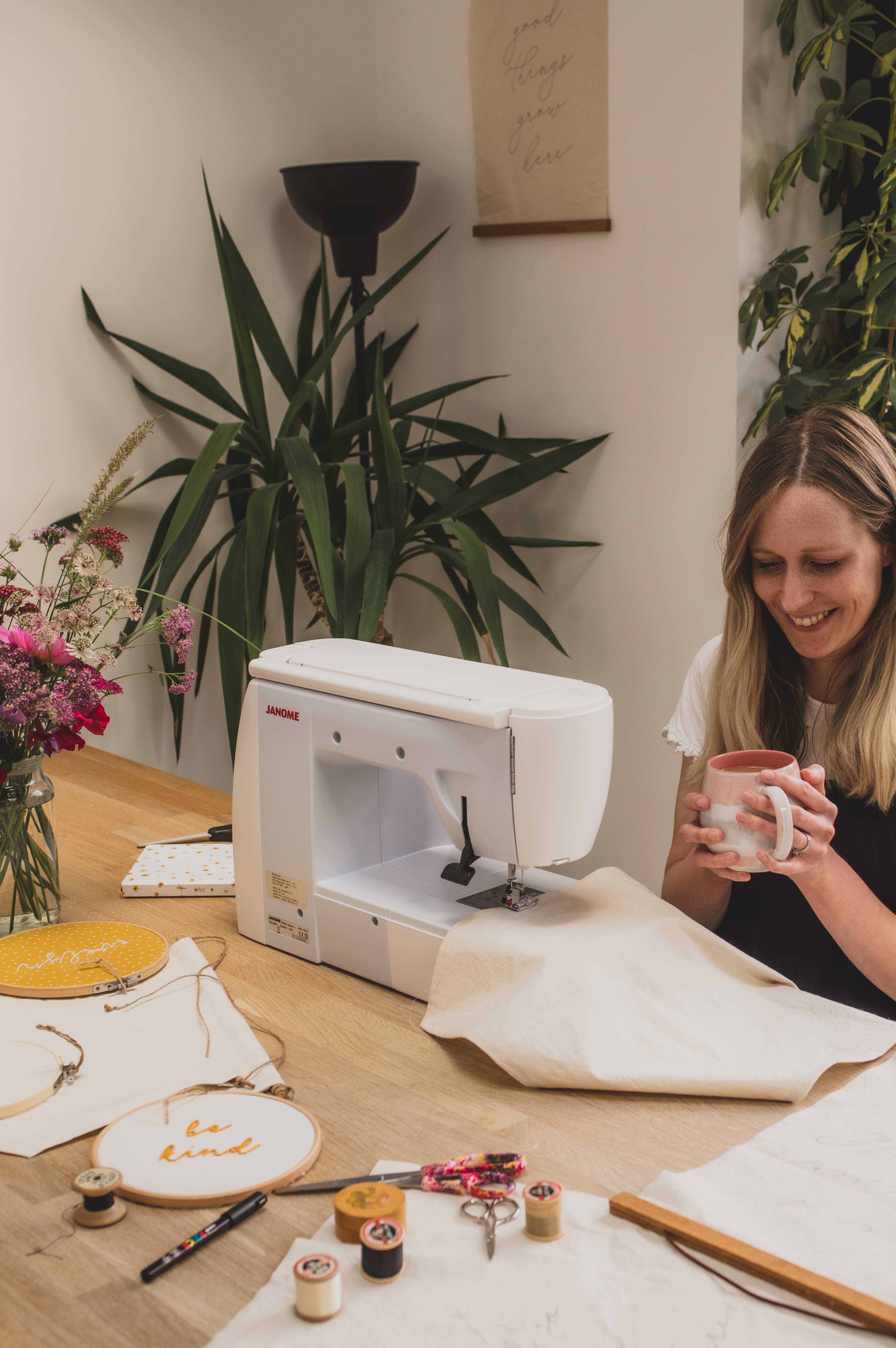 Becky sitting at table with cup of tea and sewing machine. Fabric banners and embroideries are on the table.