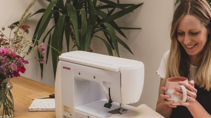 Becky sitting at table with cup of tea and sewing machine. Fabric banners and embroideries are on the table.