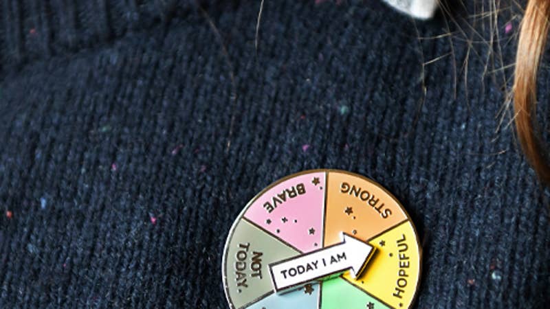 A spinning enamel pin badge, with six segments each with positive words in, is pinned to someones navy blue jumper.