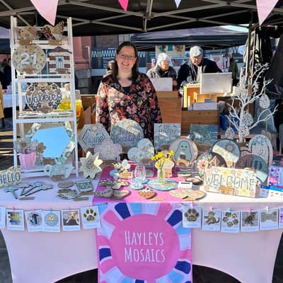 Hayley is standing behind a market stall with a pink table cloth and dark pink logo tablecloth with pretty mosaic items on display