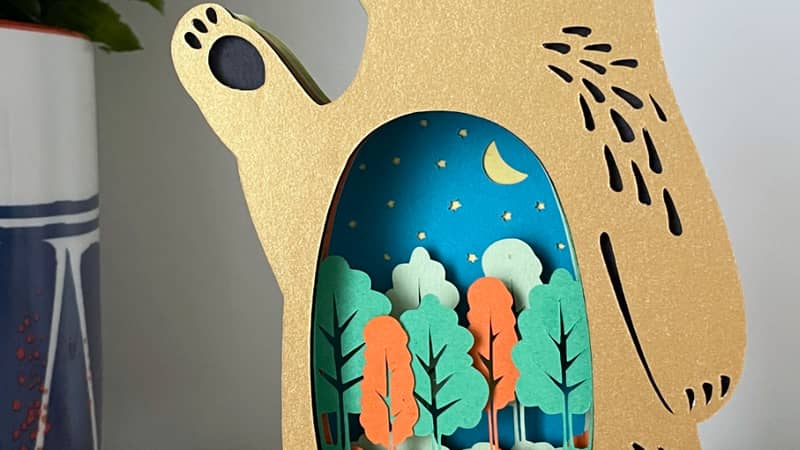 "Forest Friends" bear papercut in gold as a shelf ornament. Cut out section features a moonlit forest scene.