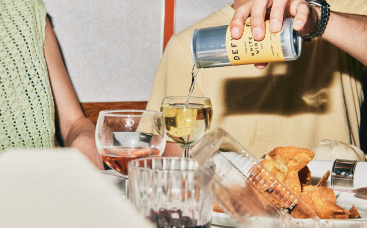 A person pouring a can of DEFY organic white wine into a glass, at a dinner table.