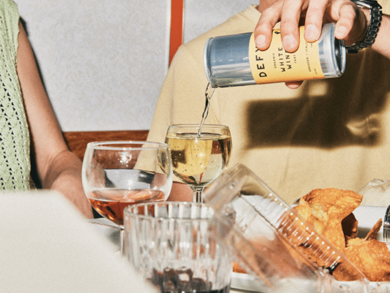A person pouring a can of DEFY organic white wine into a glass, at a dinner table.