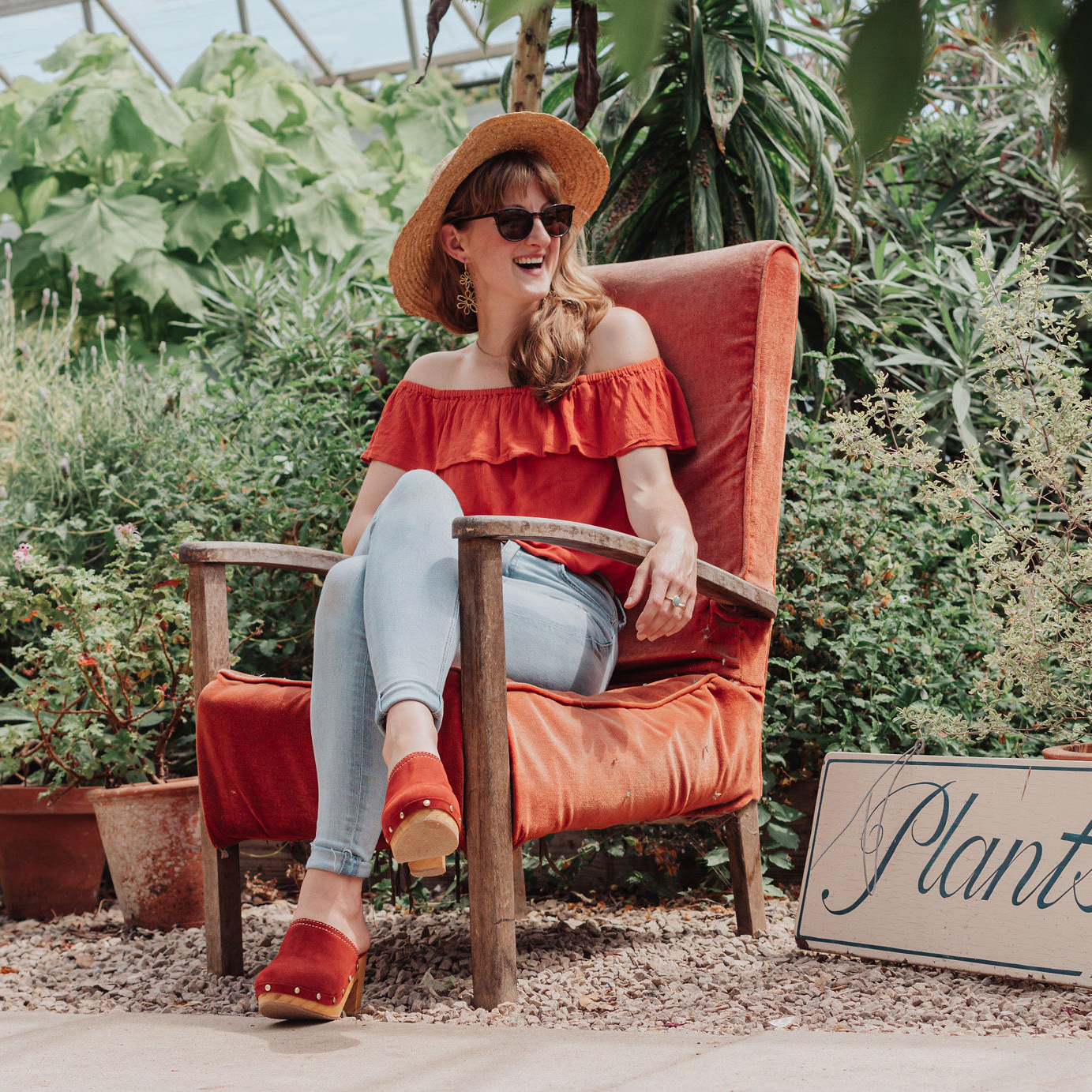 Nic laughs, posed on a photoshoot in an old orange velvet chair in a greenhouse surrounded by potted and tropical plants. 