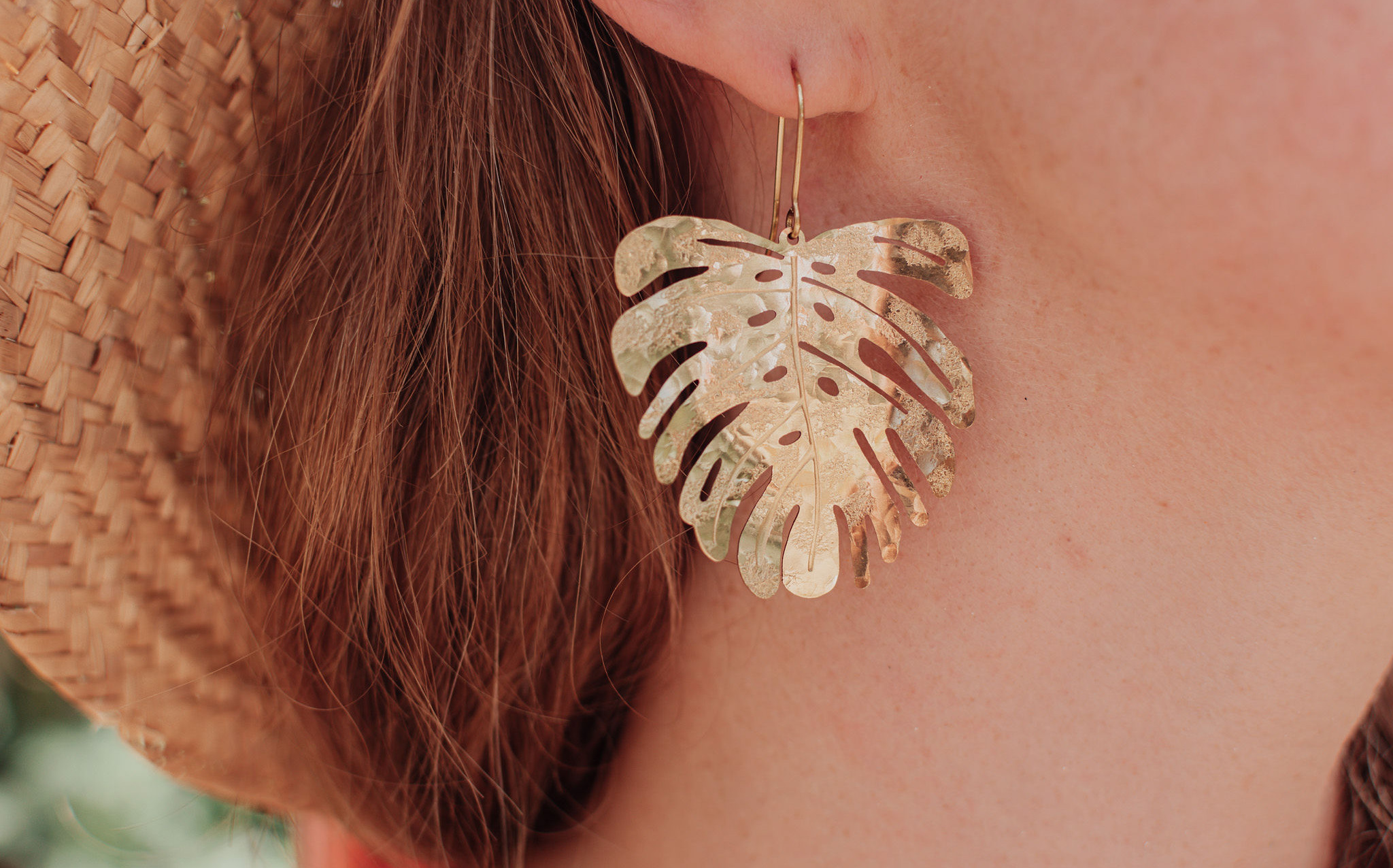 Nic wears a floppy straw hat and models a statement beaten brass earring in a Monstera leaf design. The brass Monstera leaf earring sparkles in soft glowing sun