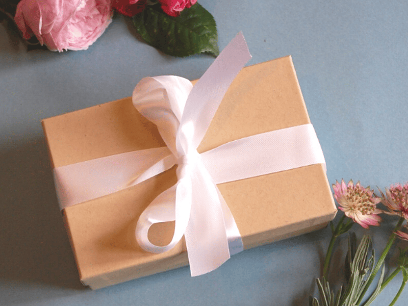 an image of a gift box tied with white ribbon next to some flowers