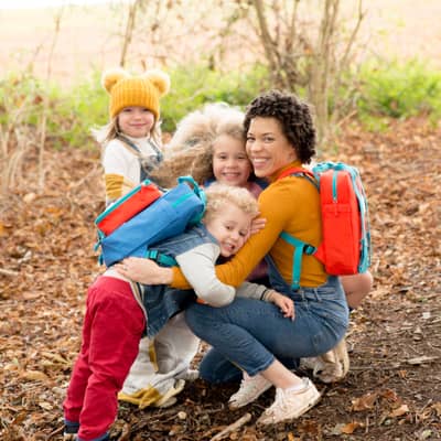 A mixed race lady cuddling three small children. They are all wearing magnetic backpacks