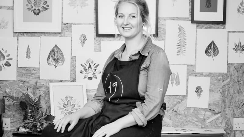 Printmaker Natalie Hallett sitting in her studio based in Devon in front of her working wall of botanical prints