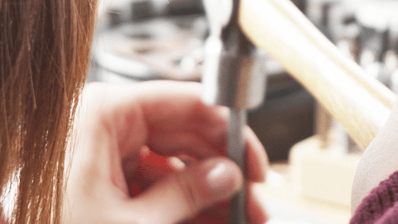 woman with brown hair at a workbench. Hammering onto a steel block