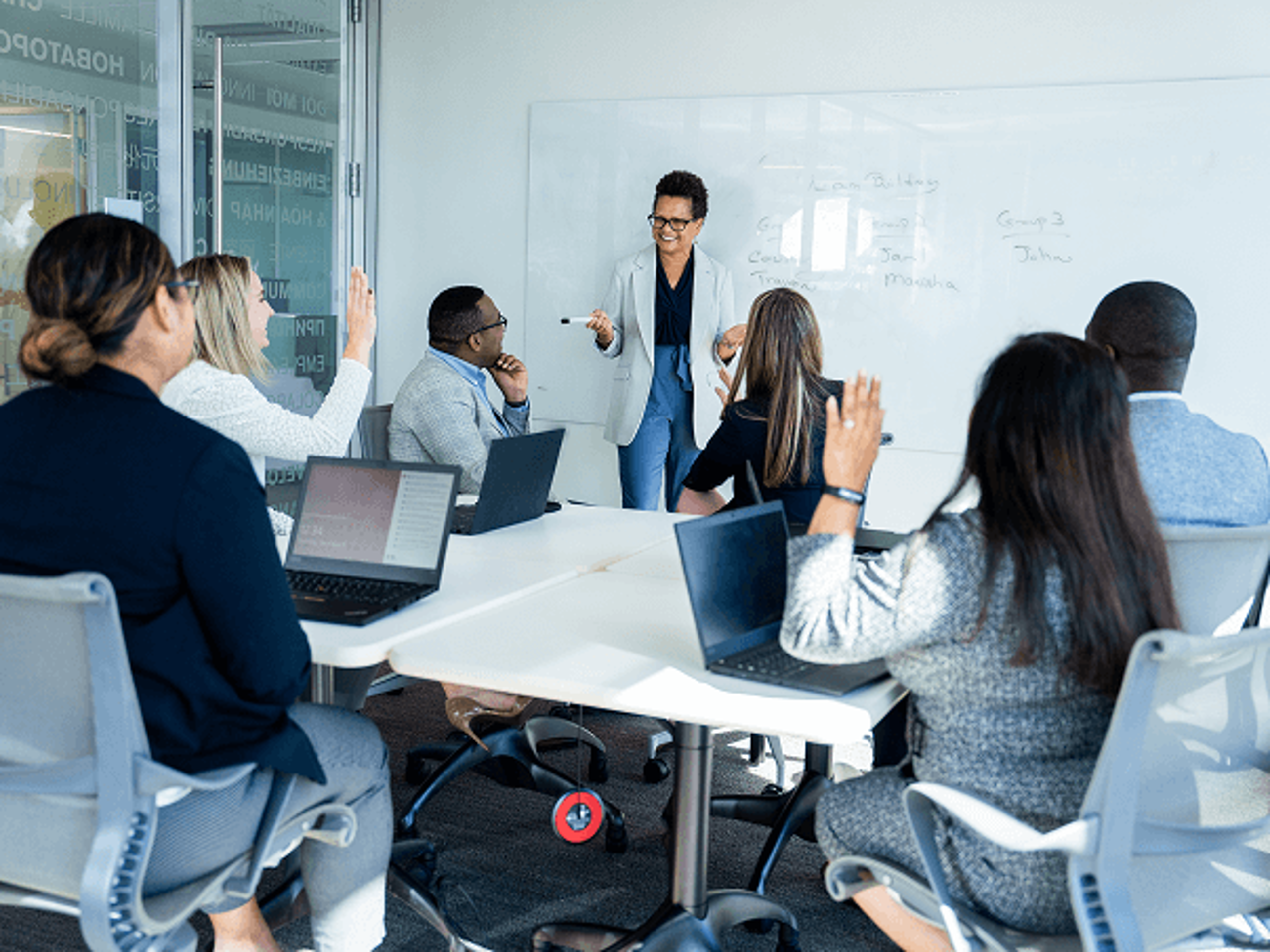 Presenter leading a meeting at a whiteboard while team members seated around a conference table raise their hands and use laptops.