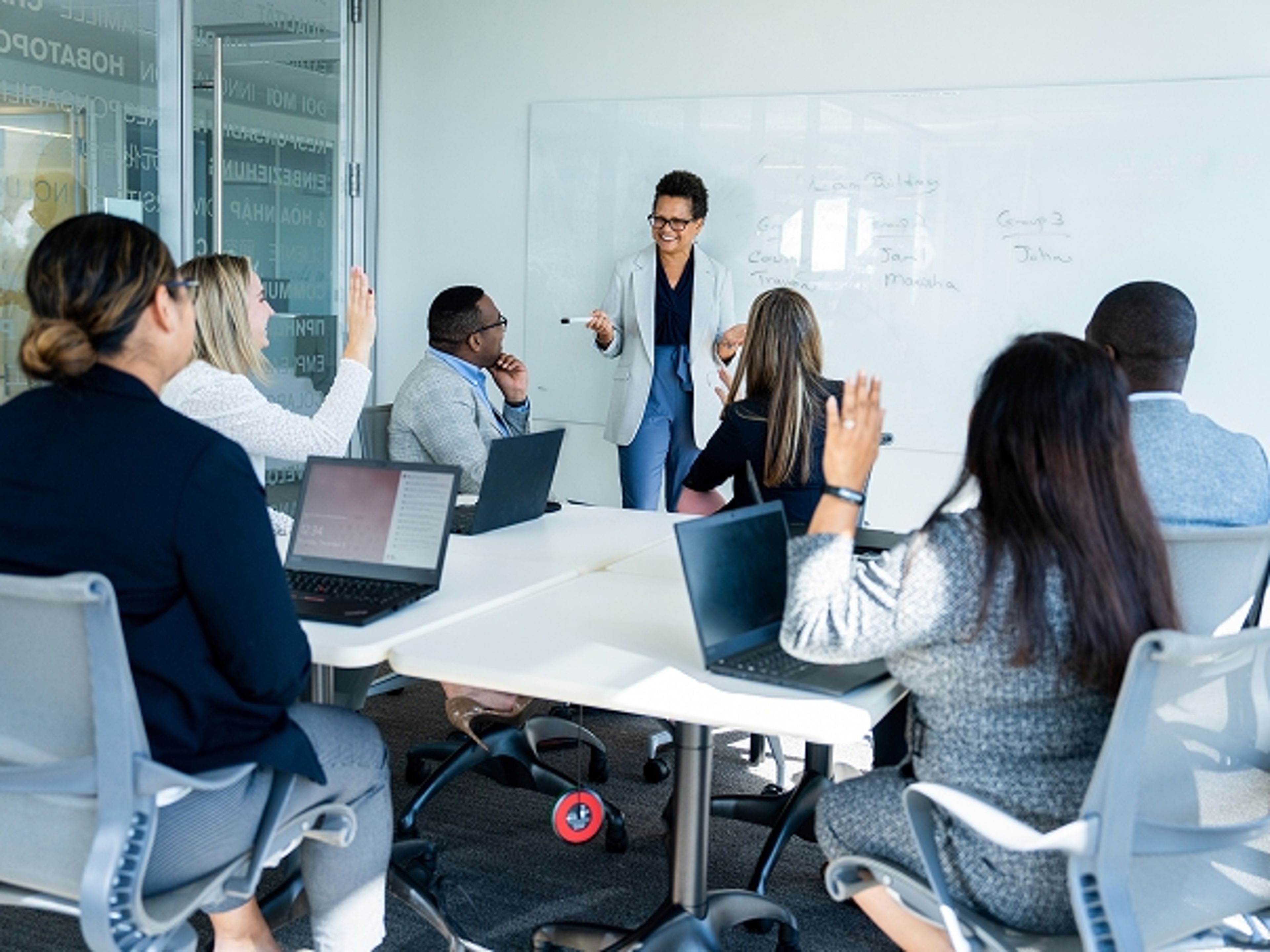Presenter leading a meeting at a whiteboard while team members seated around a conference table raise their hands and use laptops.