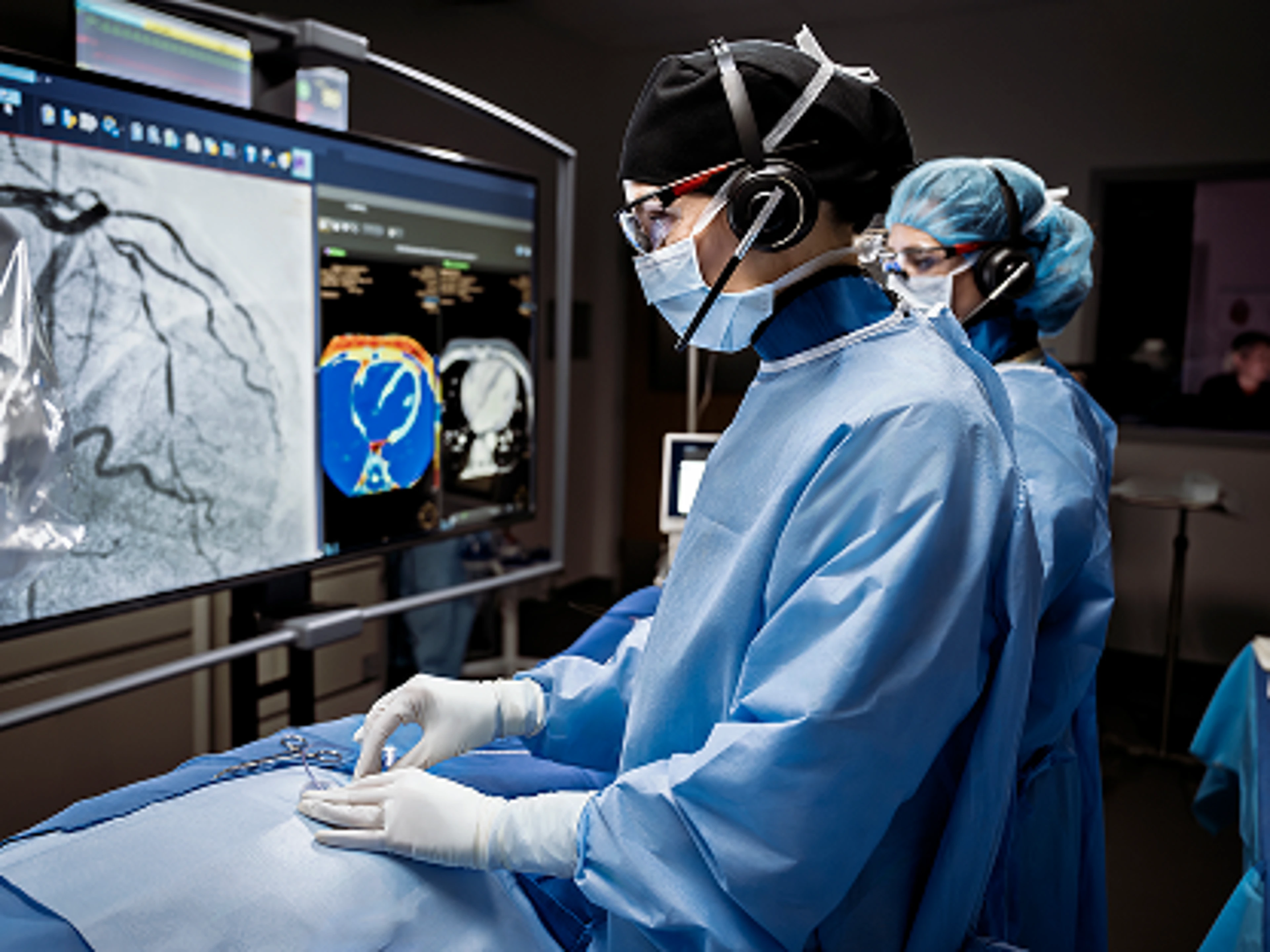 Clinicians in the cath lab perform an endovascular procedure using an Impella heart pump, with the control room team visible in the background monitoring the case.