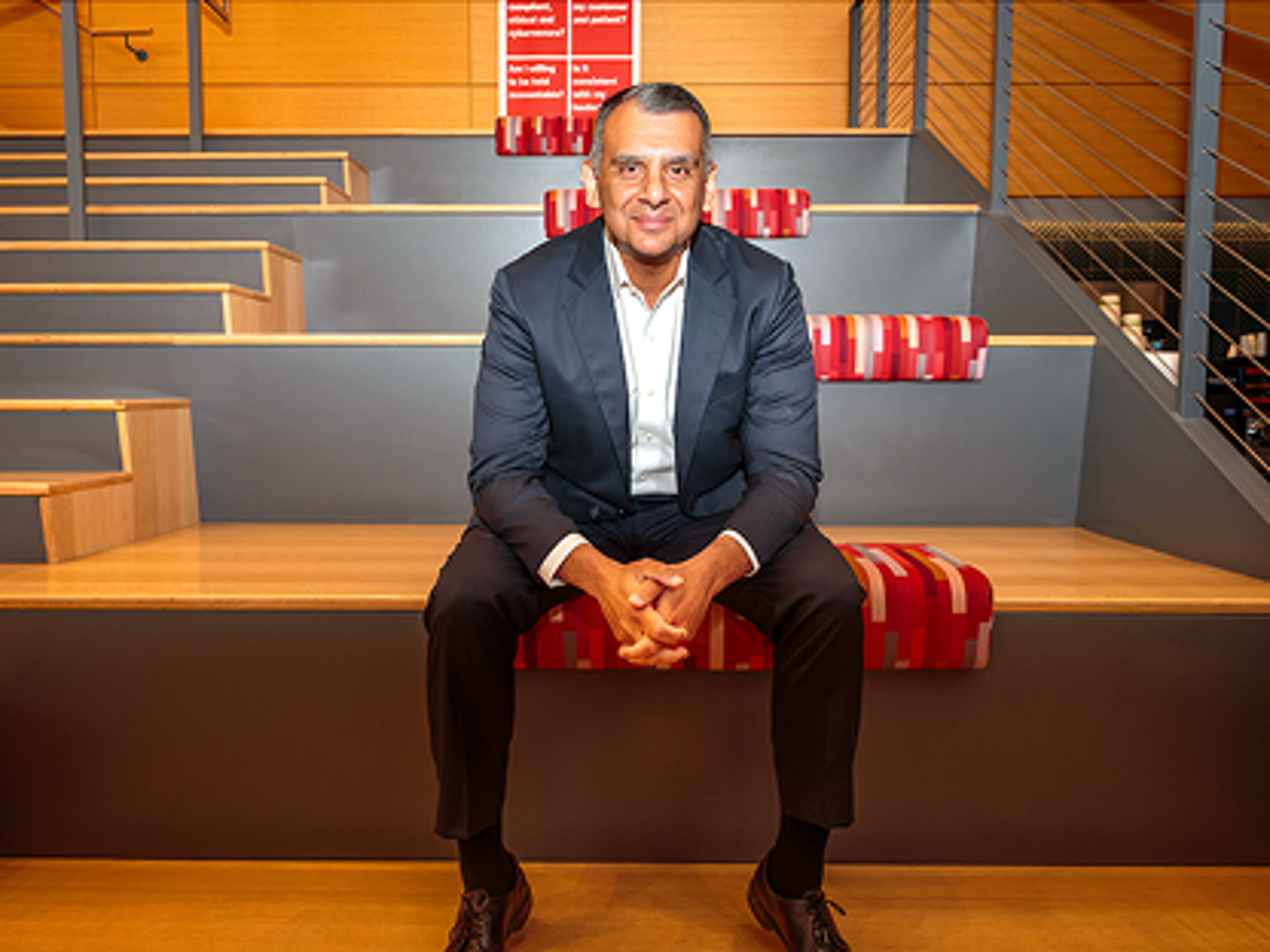 Dr. Sanjeev Aggarwal in a dark suit seated on tiered wooden steps in a modern auditorium setting with hands folded.