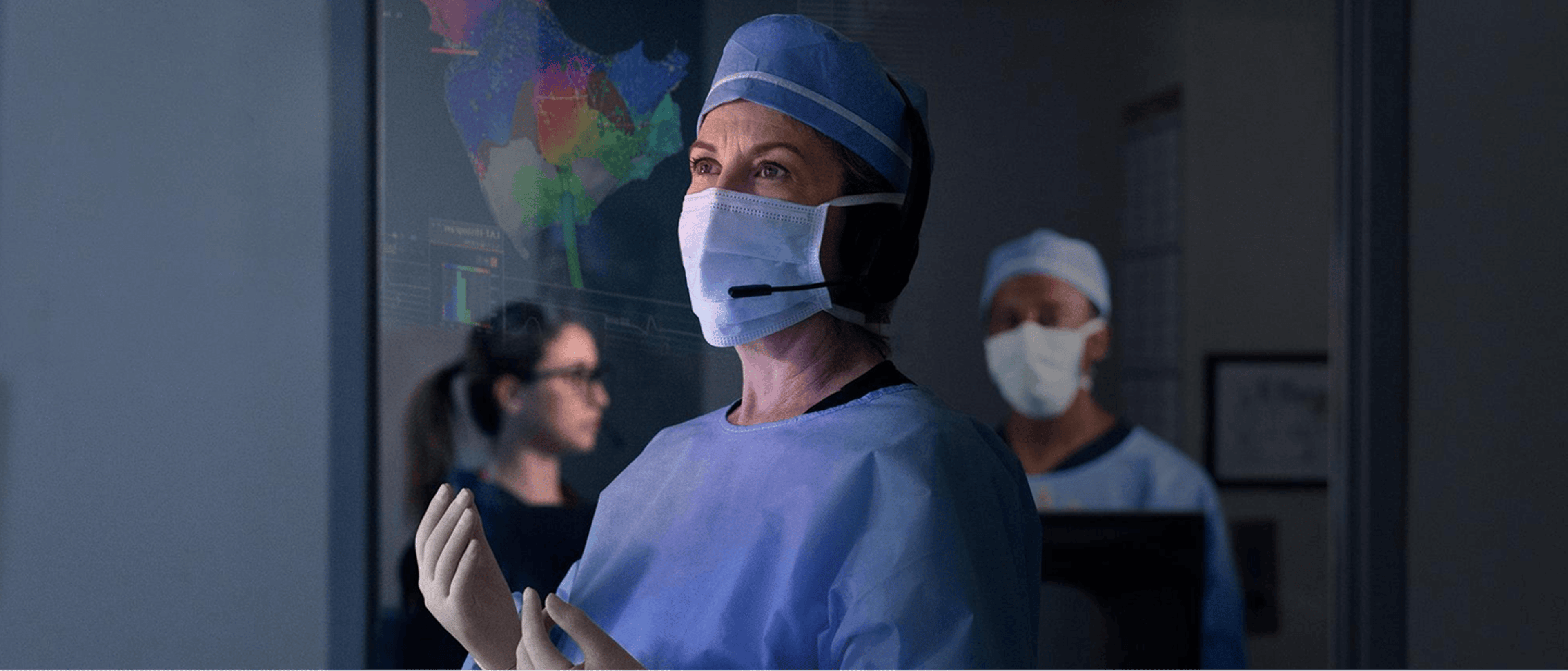 A surgeon wearing a mask, headset, and surgical scrubs stands in an operating room, looking toward a large digital display with medical imaging, while other medical staff work in the background.