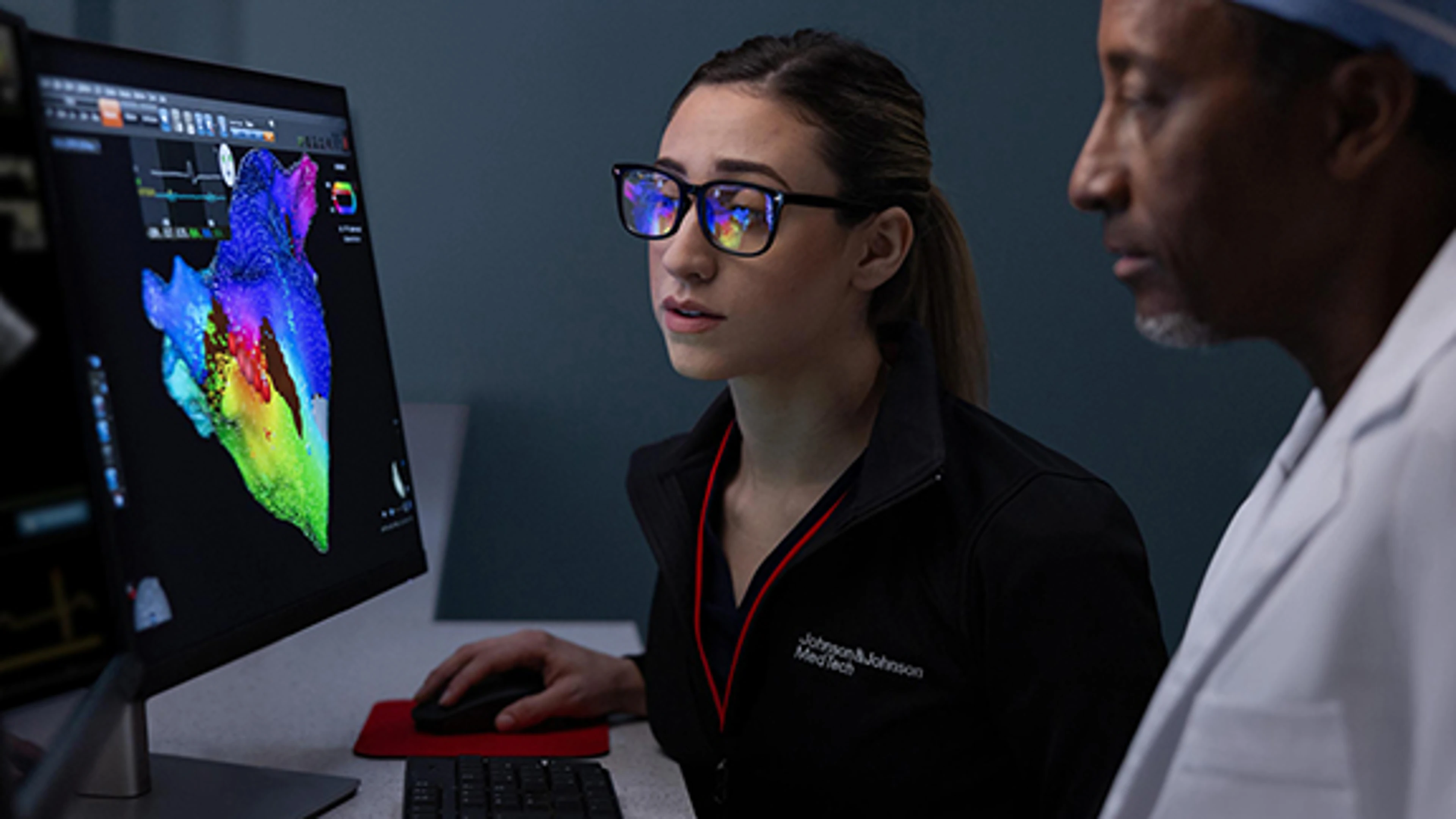 A clinical account specialist in glasses reviews a brightly colored 3D heart mapping visualization on a computer screen. A physician stands next to her as they analyze the data together.