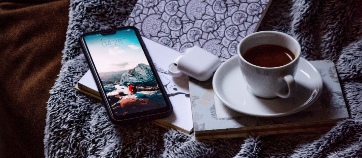 A cup of coffee, smartphone, journal and earbuds rest on top of a fuzzy blanket