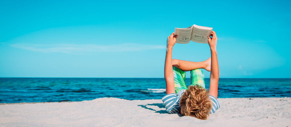 A kid lays on his back on the beach and holds up a book to read