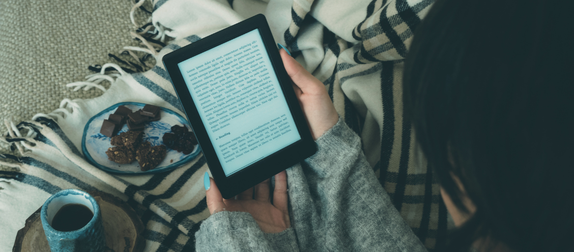 A person reads an ebook on a cozy blanket with a plate of cookies and cup of coffee.