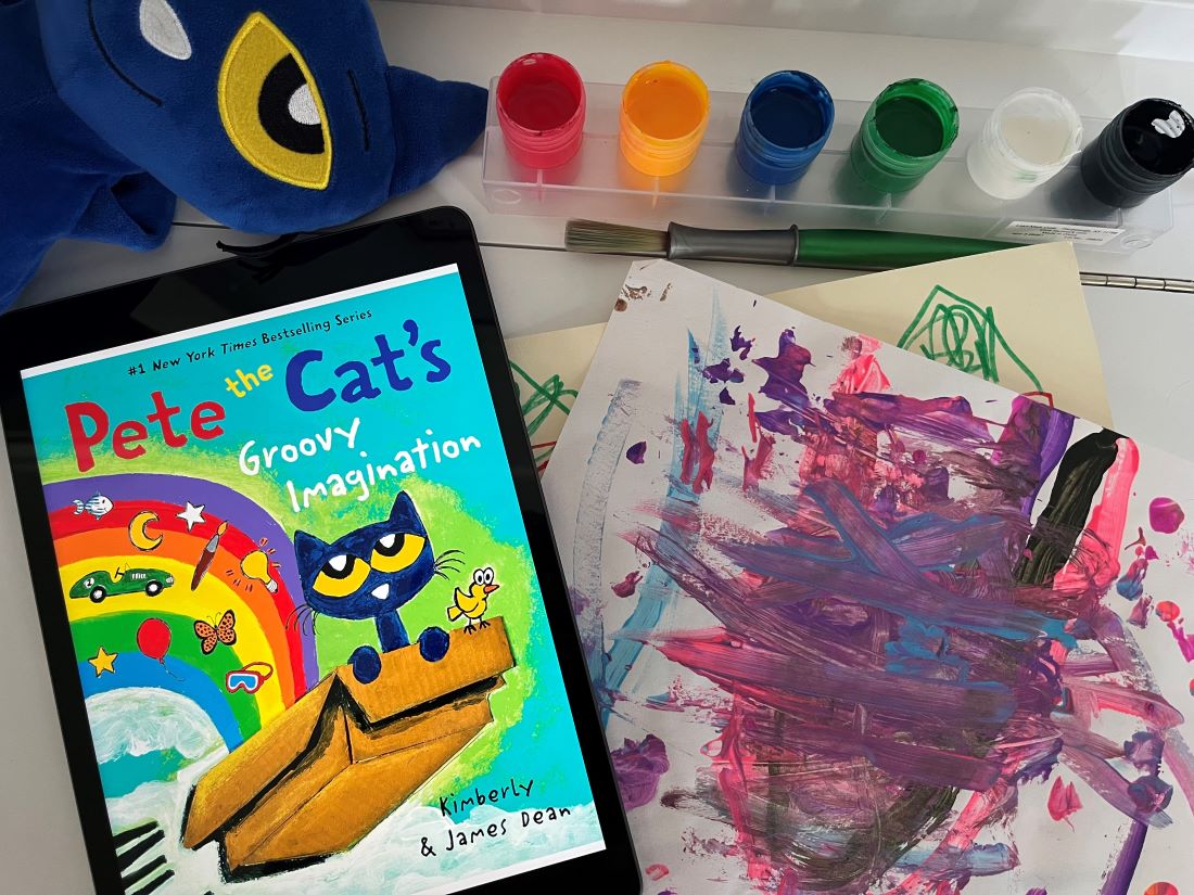 An iPad on a table with the cover "Pete the Cat's Groovy Imagination", a Pete the Cat stuffed animal, paint supplies and a child's painting.