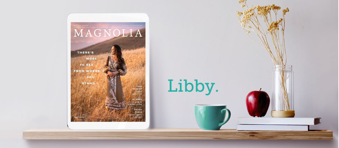 The digital magazine "Magnolia" is displayed on a tablet on a shelf next to an apple, mug, books and vase.