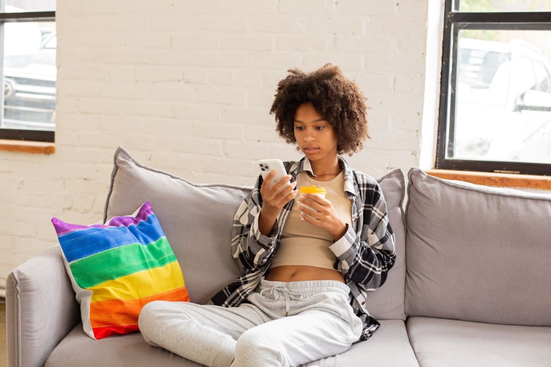 Person sitting on the couch looking at a phone next to a rainbow pillow