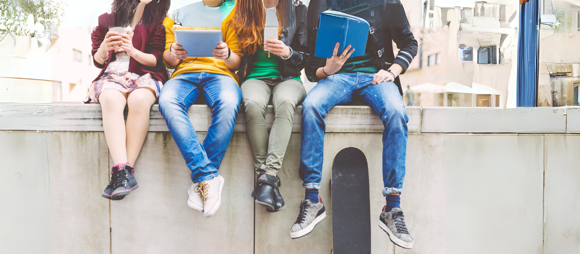 Teens sit on a ledge holding books, phones and tablets.