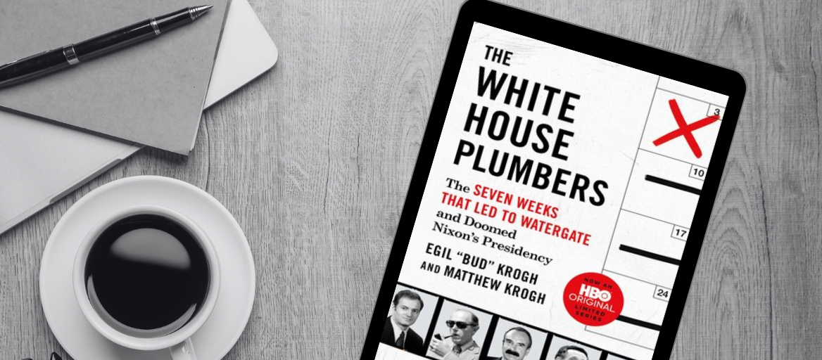 A tablet featuring the book "The White House Plumbers" sits on a desk next to a cup of coffee, pen and notebook