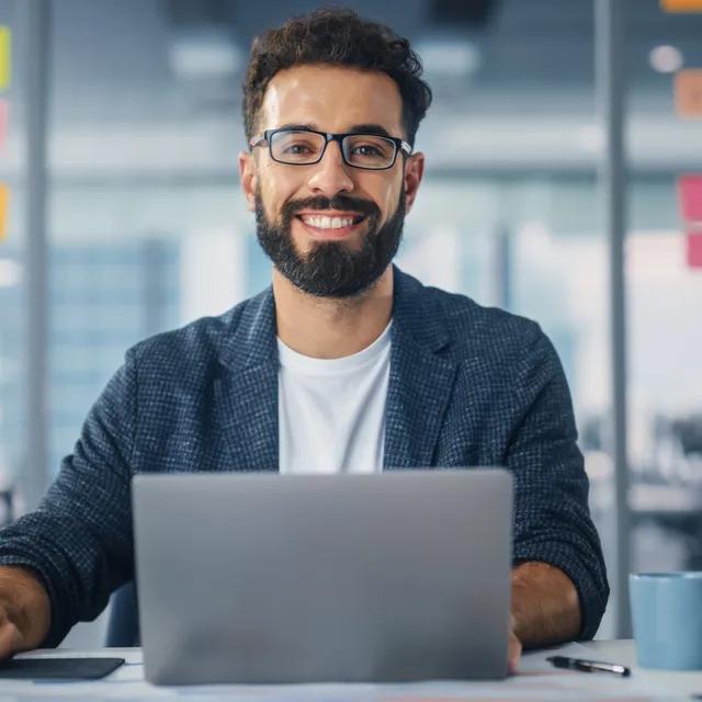 Hombre joven con barba y gafas sonríe mientras está sentado frente a una laptop.