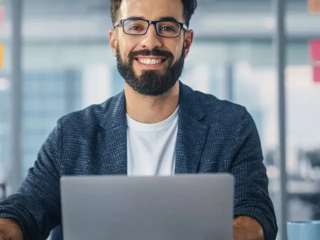 Hombre joven con barba y gafas sonríe mientras está sentado frente a una laptop.