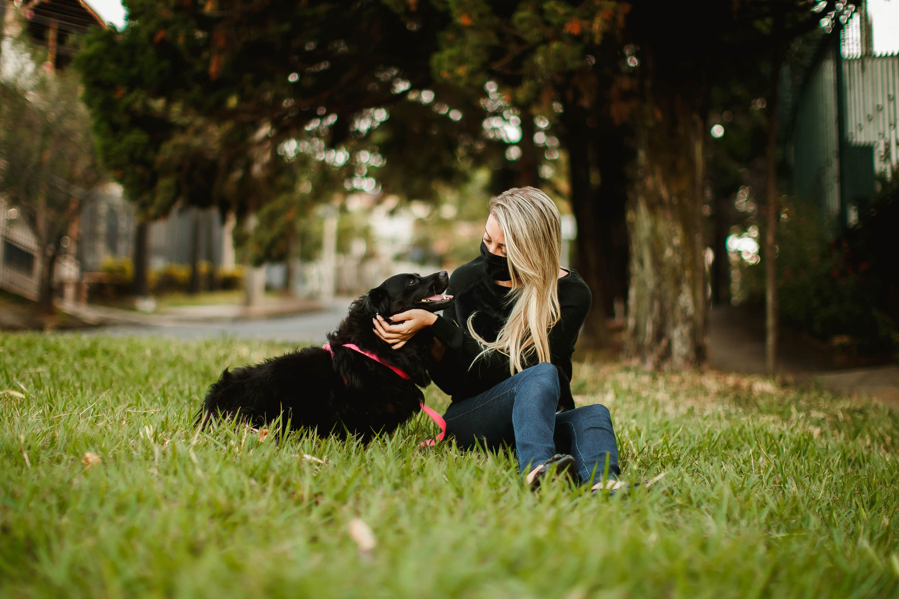 woman in mask sitting with black dog in grass sniffing her face