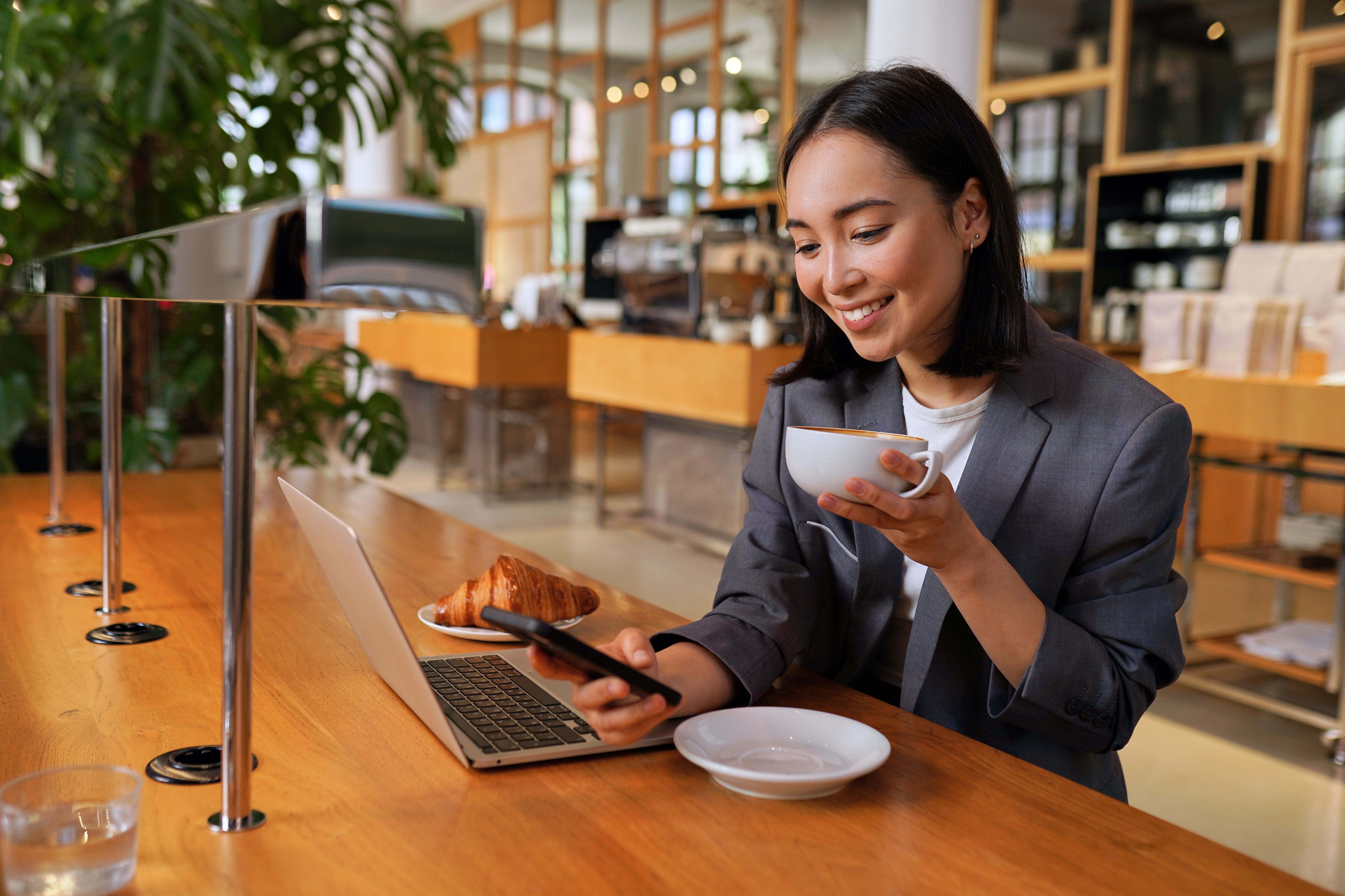 Woman sitting at a coffee shop drinking coffee holding her phone and doing a self-paced language learning course with Berlitz on her laptop