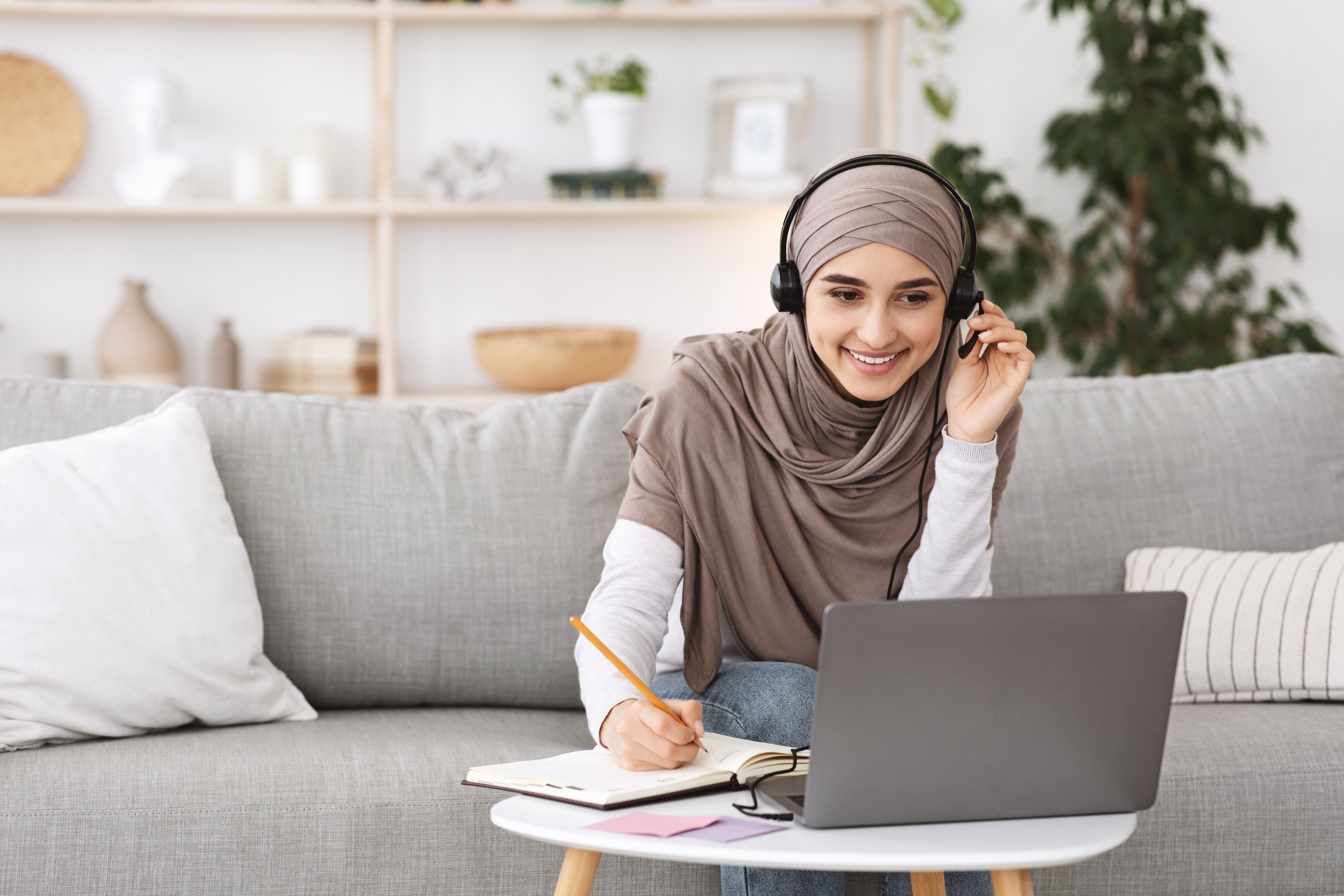 Woman in a scarf and with headphones making notes during a private online language course she's attending from her laptop