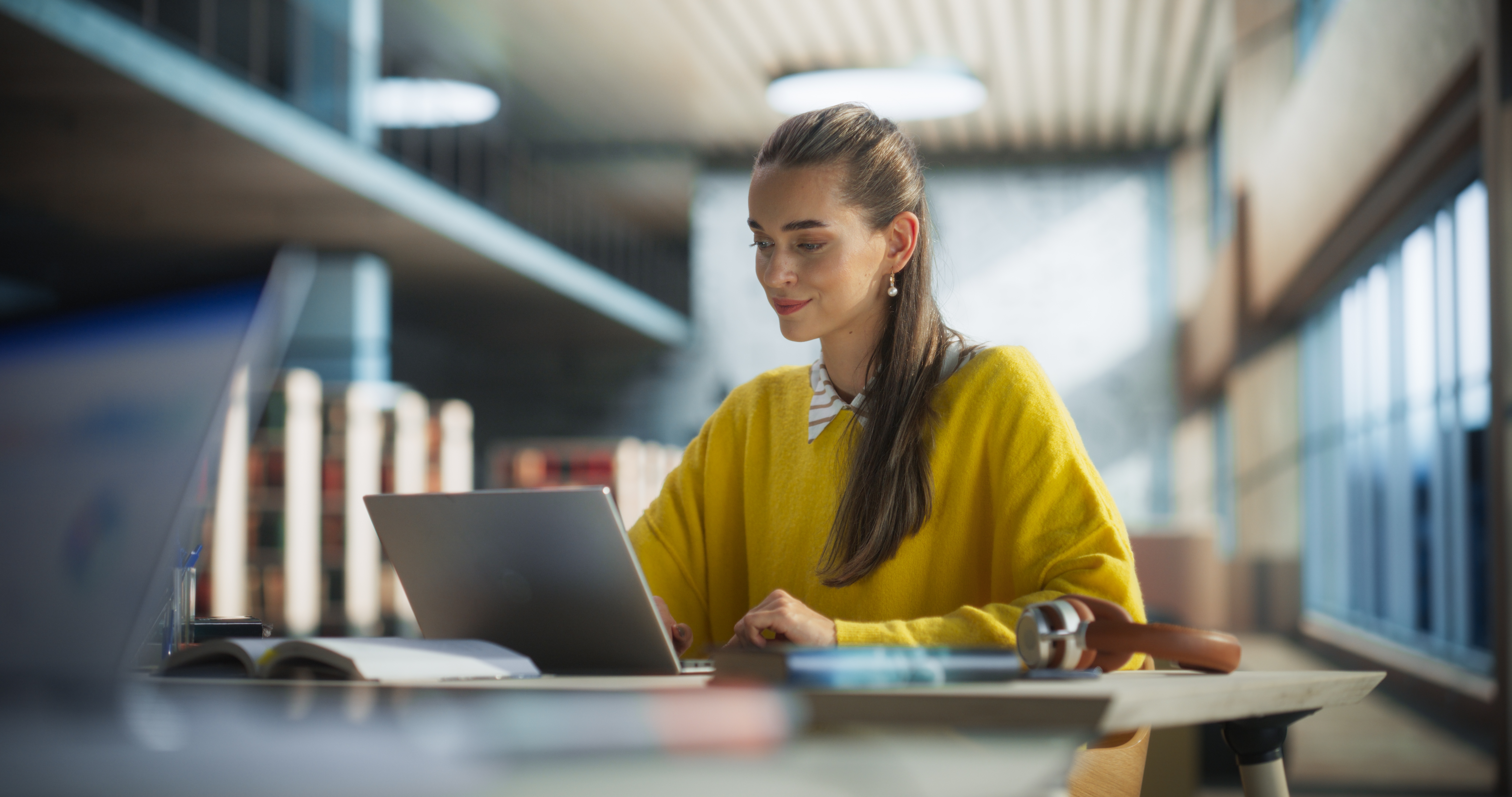 Woman in a yellow sweater sitting at a table in a library and learning a foreign language with Berlitz Flex online