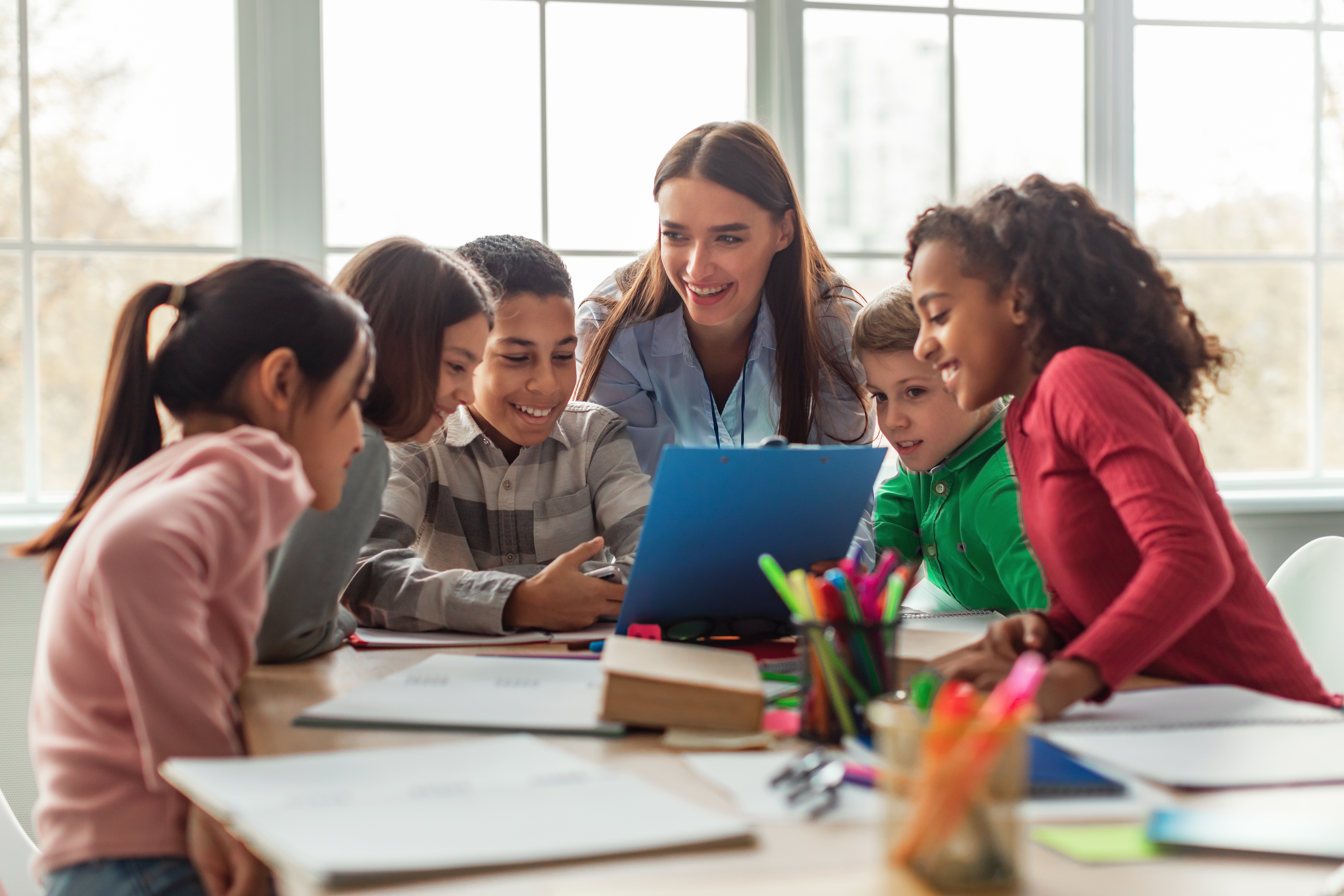 Kids sitting around a laptop and some pencils and talking to their instructor in a foreign language during Summer School