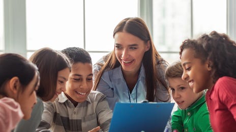 Kids sitting around a laptop and some pencils and talking to their instructor in a foreign language during Summer School