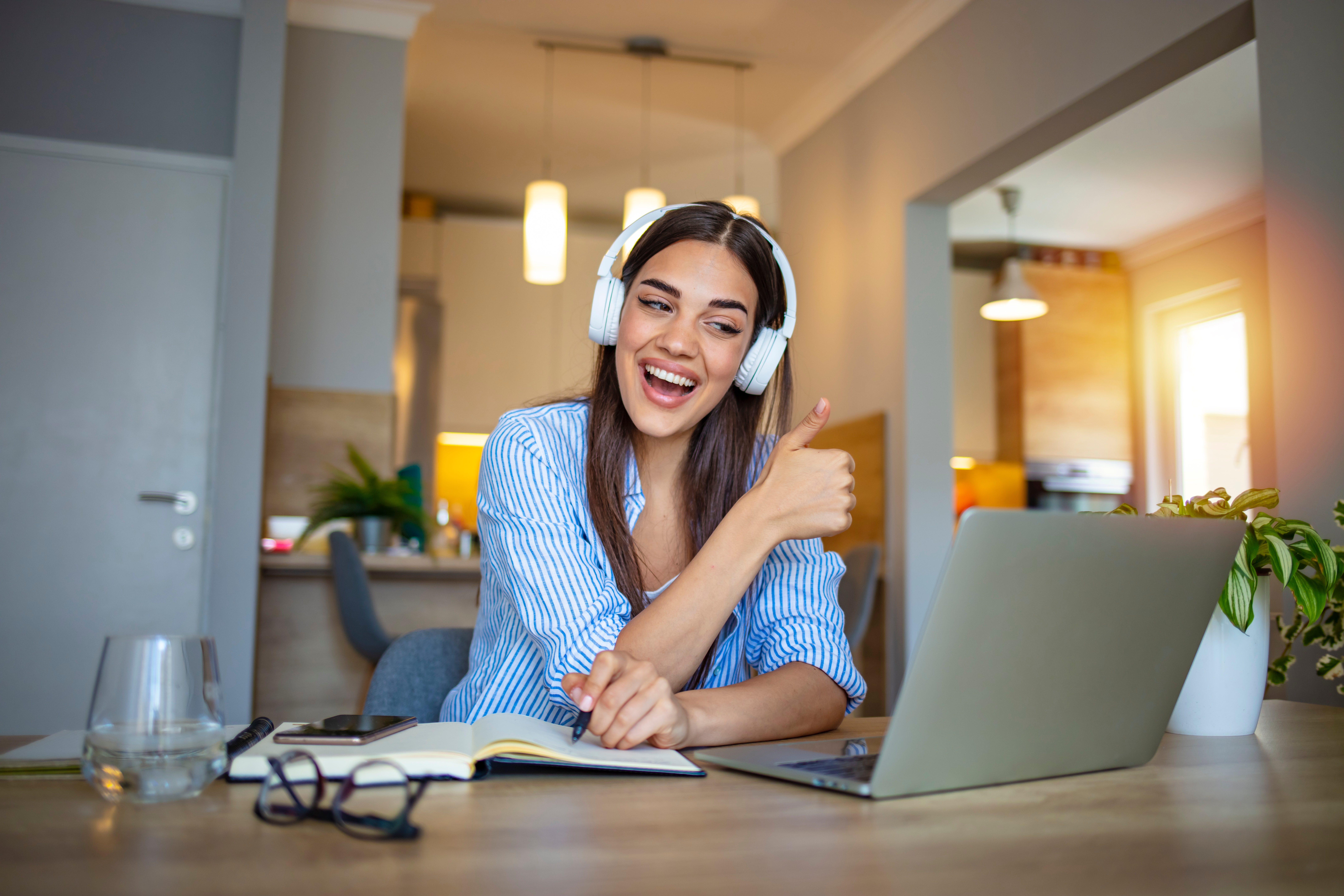 Woman with headphones holding up her thumb and smiling at her classmates during an online group language lesson she is attending from her laptop in the living room