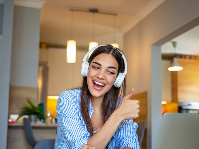 Woman with headphones holding up her thumb and smiling at her classmates during an online group language lesson she is attending from her laptop in the living room
