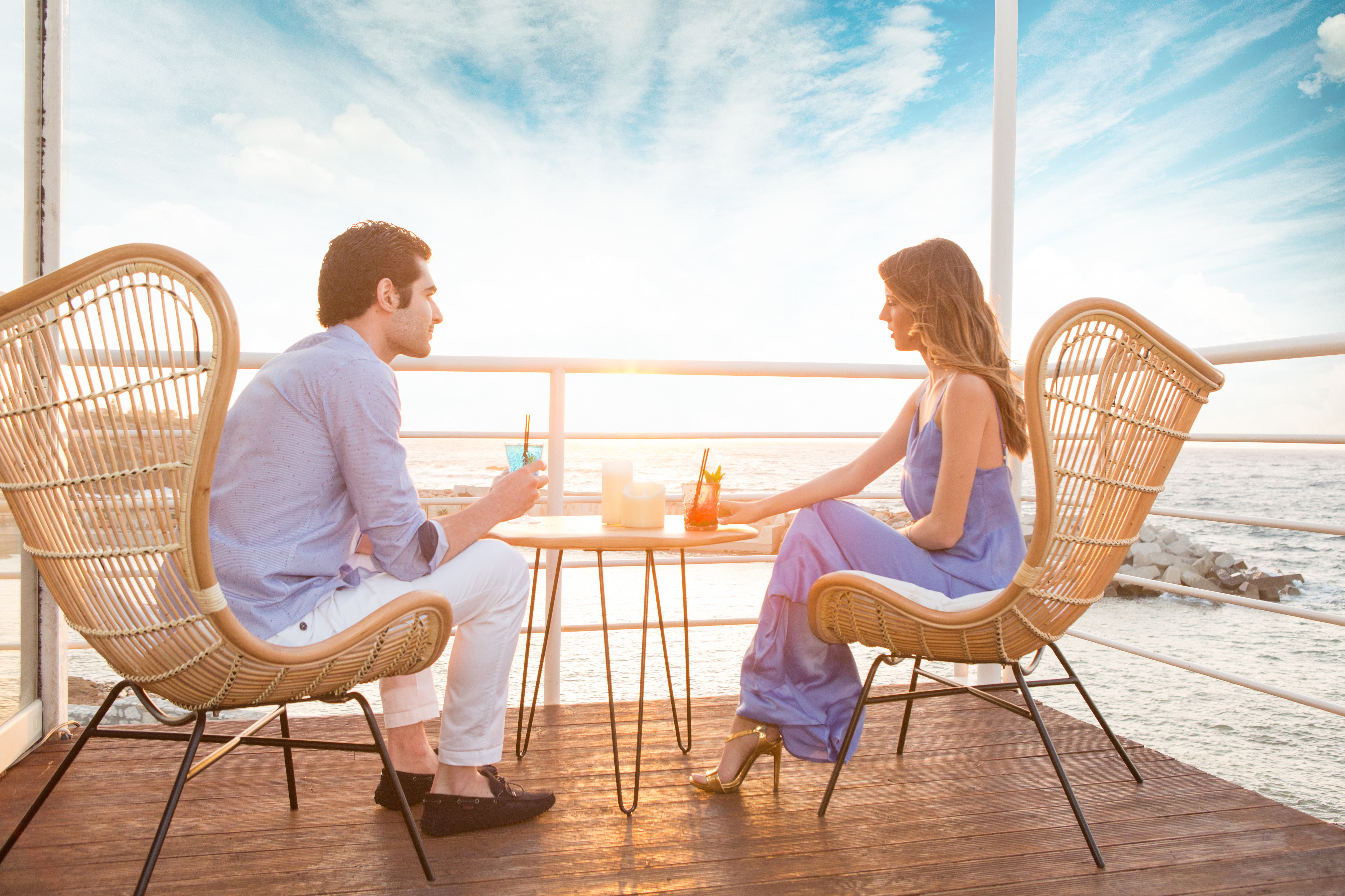 A_couple_sits_at_an_oceanfront_terrace_with_the_woman_wearing_a_flowy_maxi_dress_and_heels.jpeg