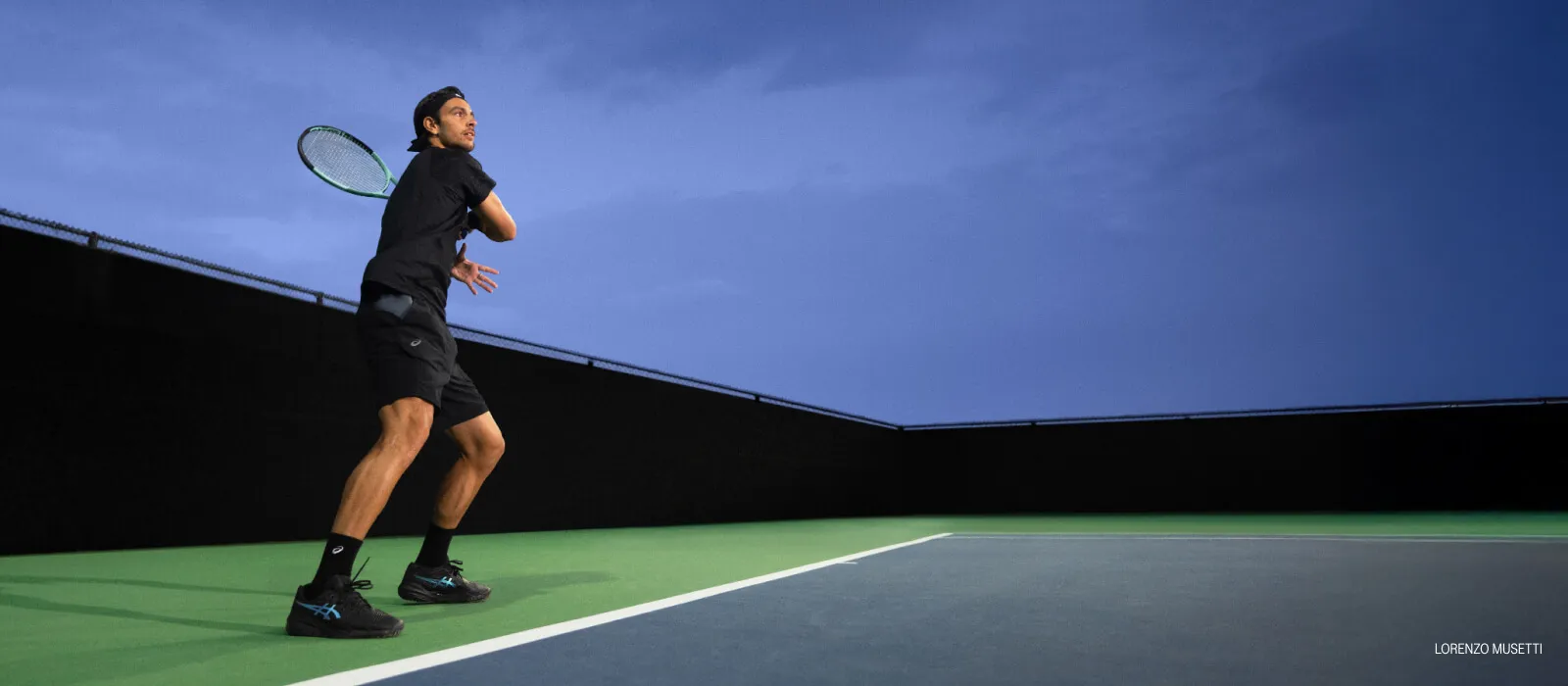 Athlete Lorenzo Musetti playing tennis wearing a black tennis clothing and black tennis shoes.