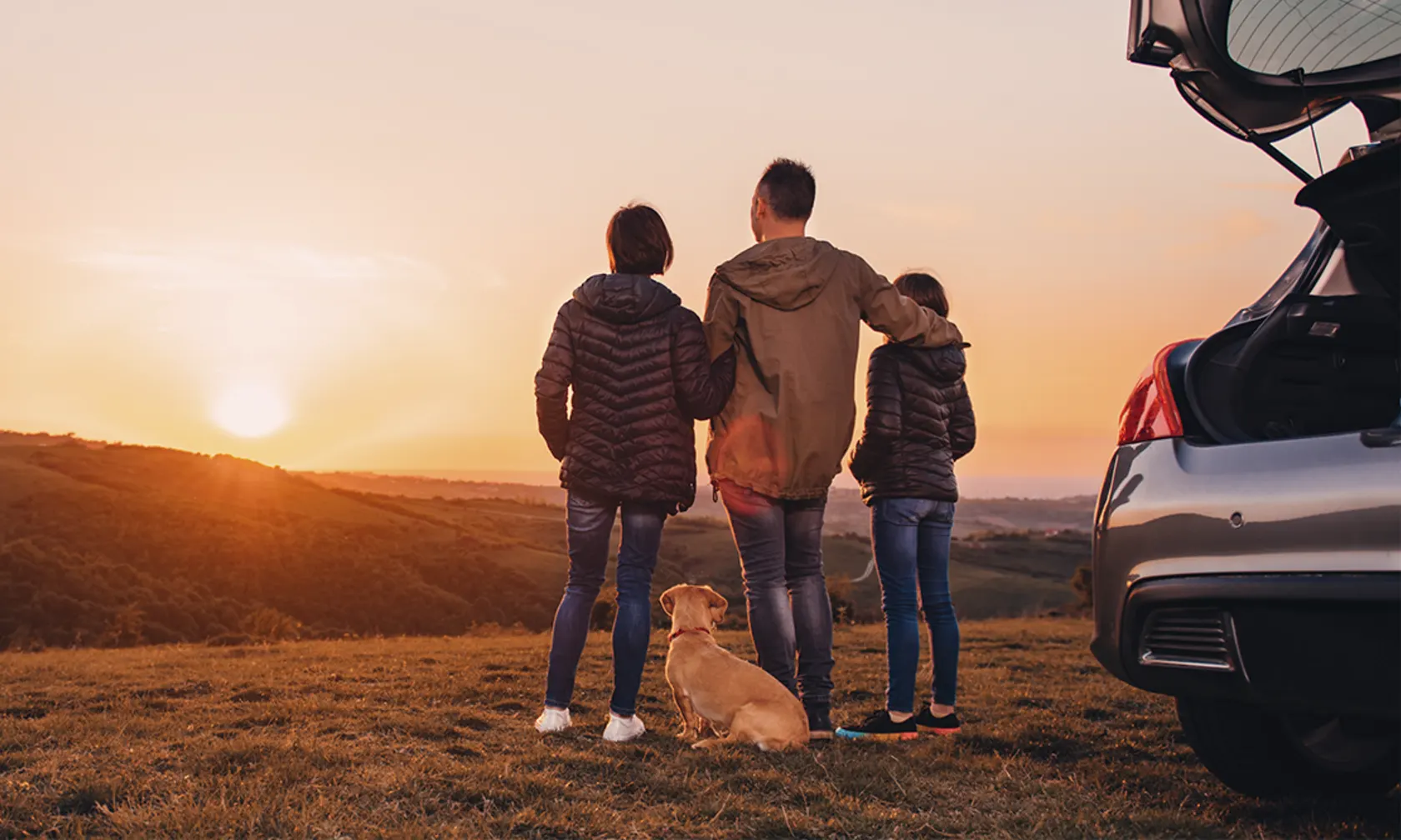 A family of three stands with their dog outside watching a sunset