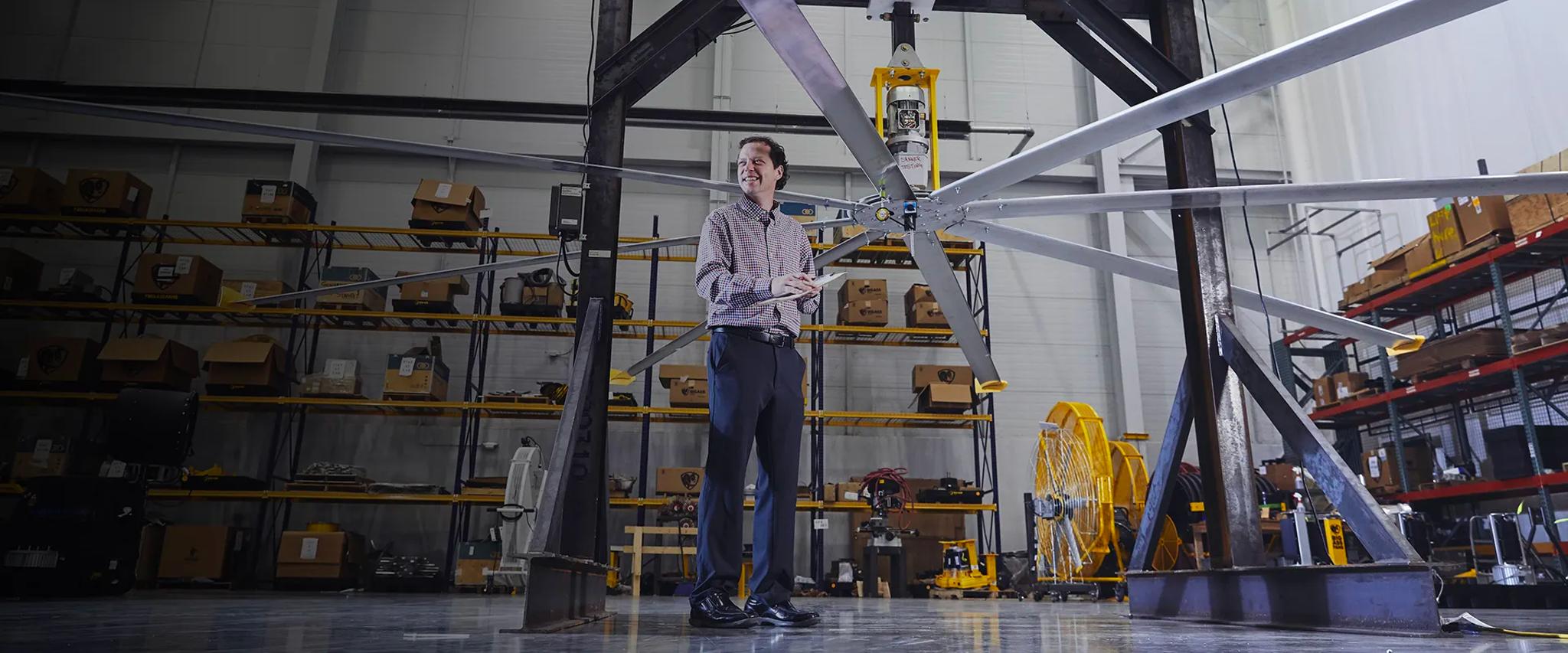 A man stands beneath a large HVLS fan in a warehouse testing facility filled with shelving and equipment