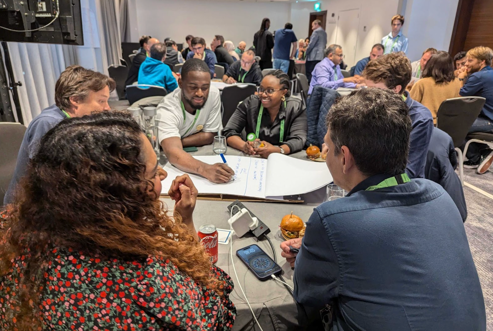 Photo of .local London AI Build Together attendees brainstorming AI solutions for the healthcare industry. The group is sitting around a table and writing down ideas on a giant notepad. The room is filled with more tables of other groups doing the same thing.