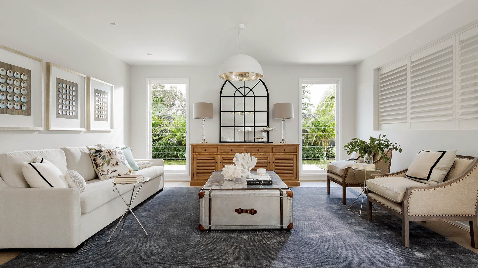 Modern living room area styled with chandelier, big arched mirror, and floor to ceiling windows.