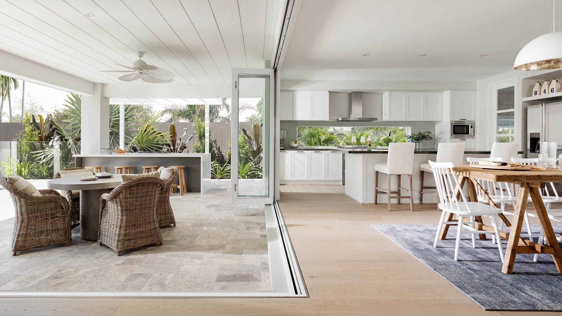 Spacious kitchen and living area connected to an alfresco, surrounded by greenery and styled with modern white furniture.