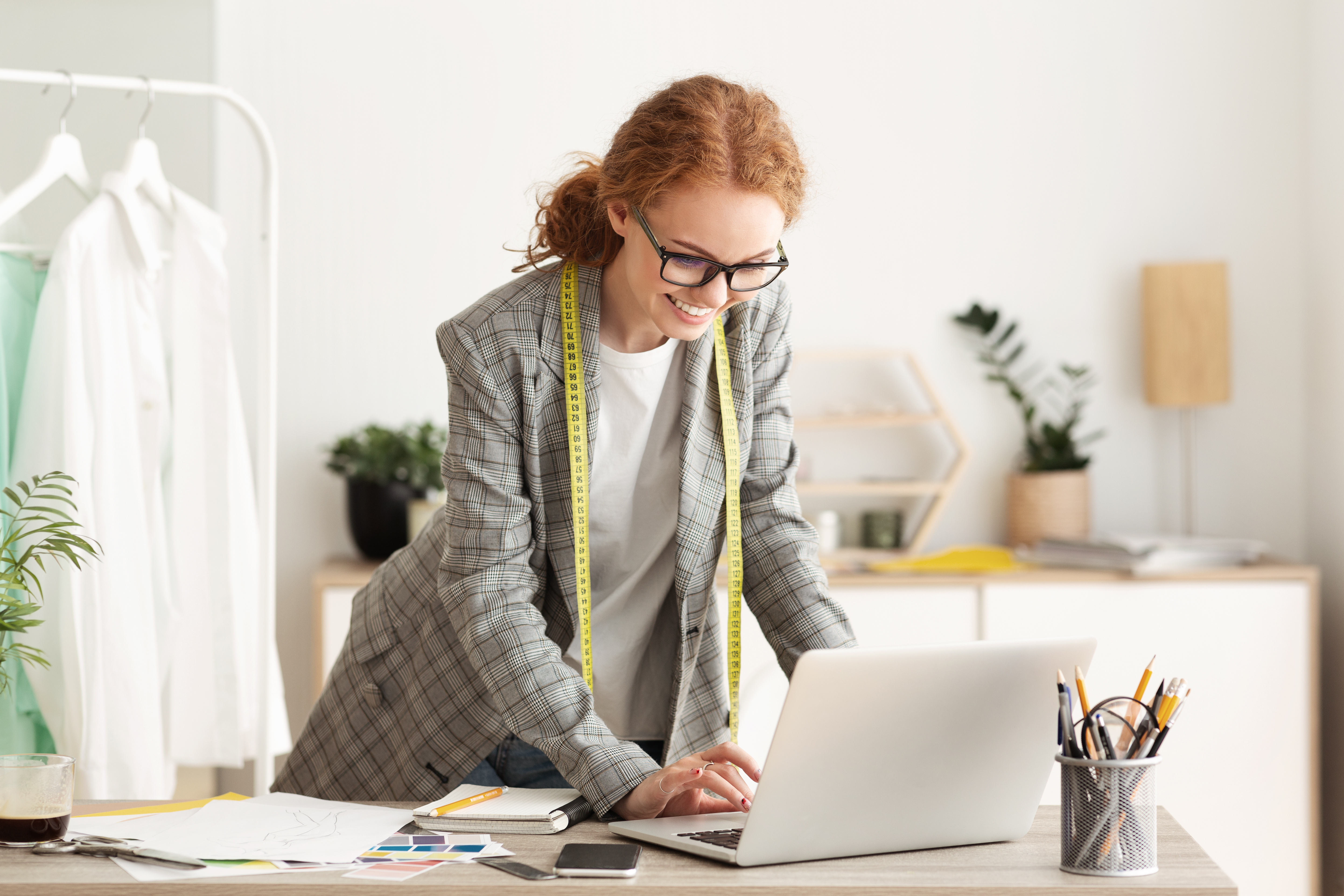 Smiling_woman_using_a_laptop_at_her_desk_with_a_tape_measure_to_check_Chicos_sizing.jpeg