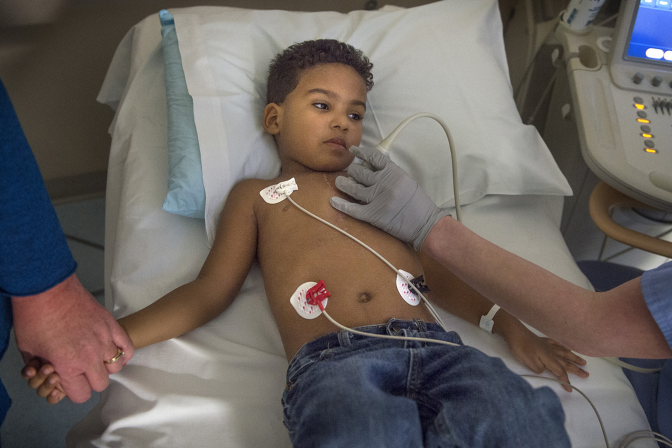 Child wearing heart monitors lying in bed while healthcare provider uses imaging device on his chest.