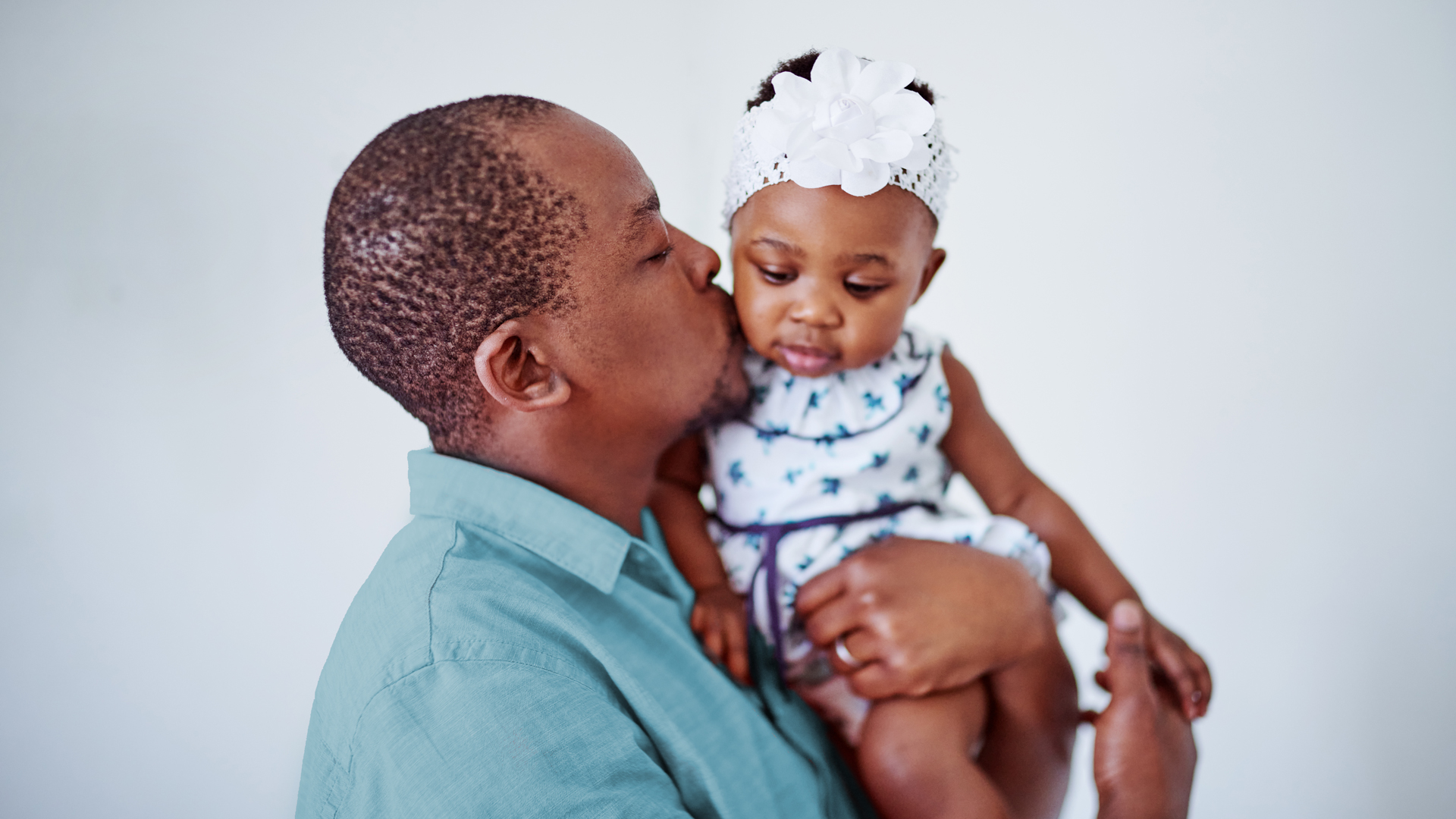 African American man in a light blue shirt holding a toddler and kissing her on the cheek