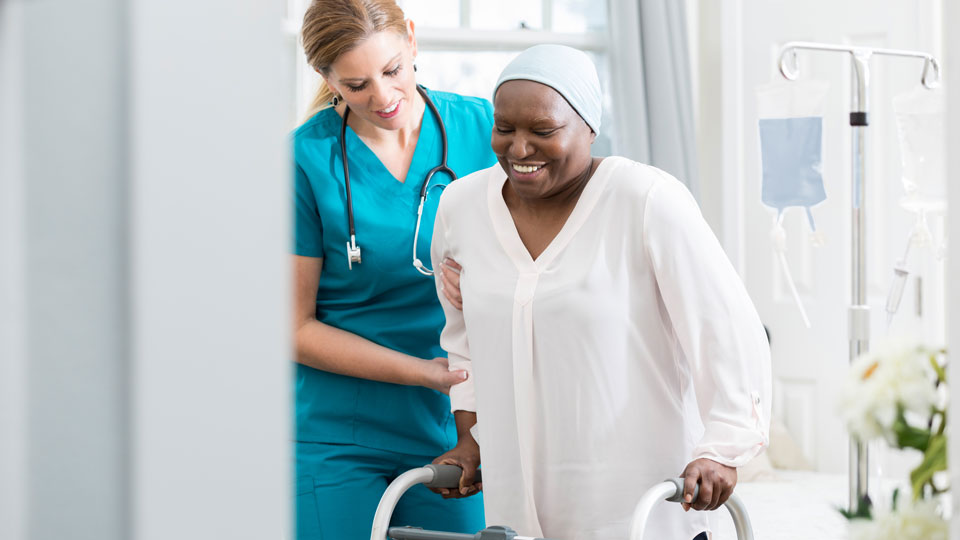 white woman in a medical uniform helping a black woman stand with a walker