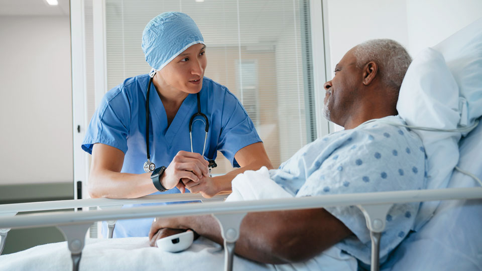 Doctor in blue scrubs with surgical cap leaning down and speaking with patient in bed