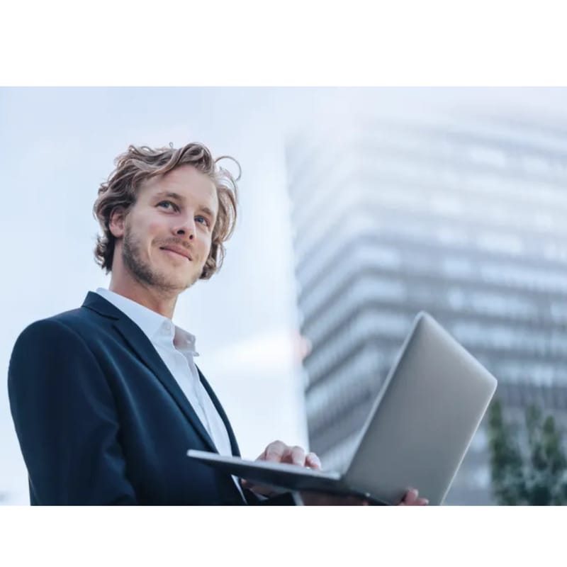 A well-dressed professional stands outdoors holding a laptop, with a modern office building in the background, suggesting work, technology, and a corporate setting.